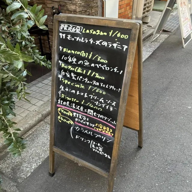 メニューが手書きされた屋外の黒板。ラザニア、10種の魚のペペロンチーノ、生うにのパスタ、リゾットなどの料理名と価格が記載されている