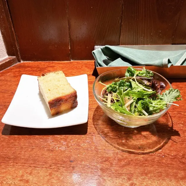 木目調のテーブルに置かれた、白い皿に乗った一切れのパン（フォカッチャ）とガラスボウルに入ったグリーンサラダ